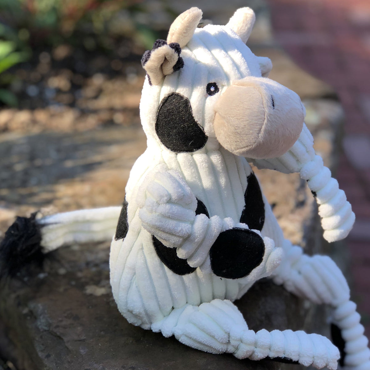 Corduroy cow Knottie sitting on a stone wall outdoors, with its tufted tail and a small bow on one ear
