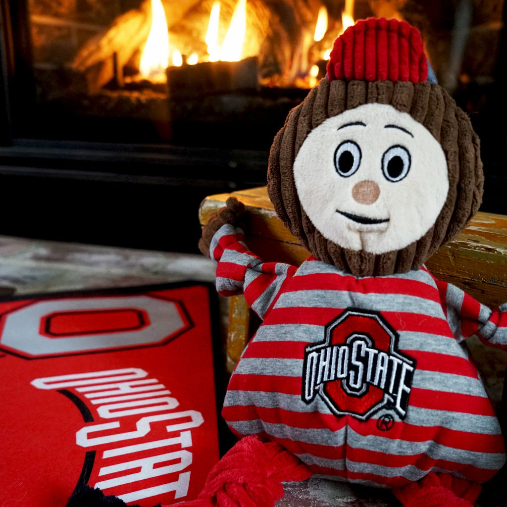 Brutus Knottie toy sitting by a lit fireplace next to an Ohio State pillow