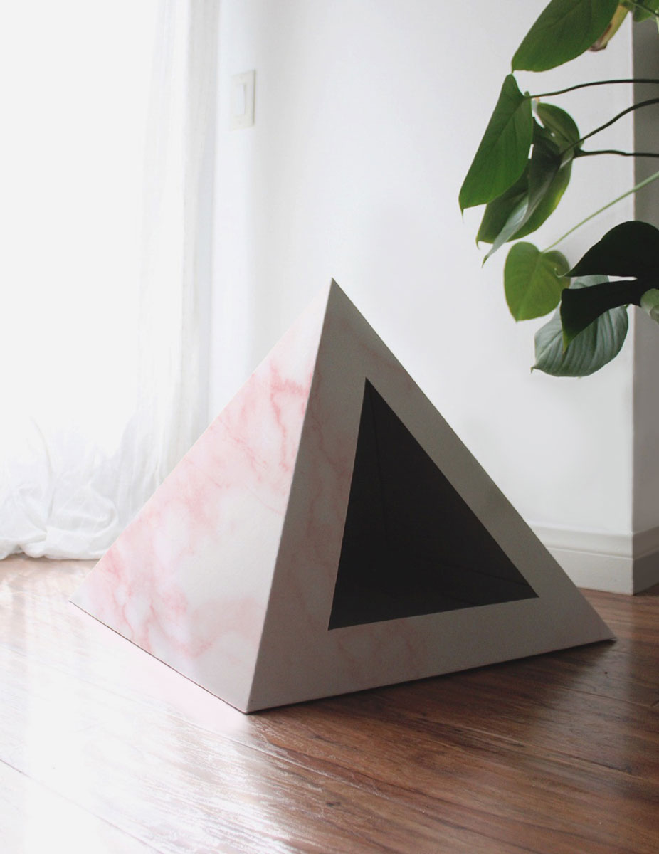 Empty pink marble-print cardboard cat pyramid on a wood floor beside a plant