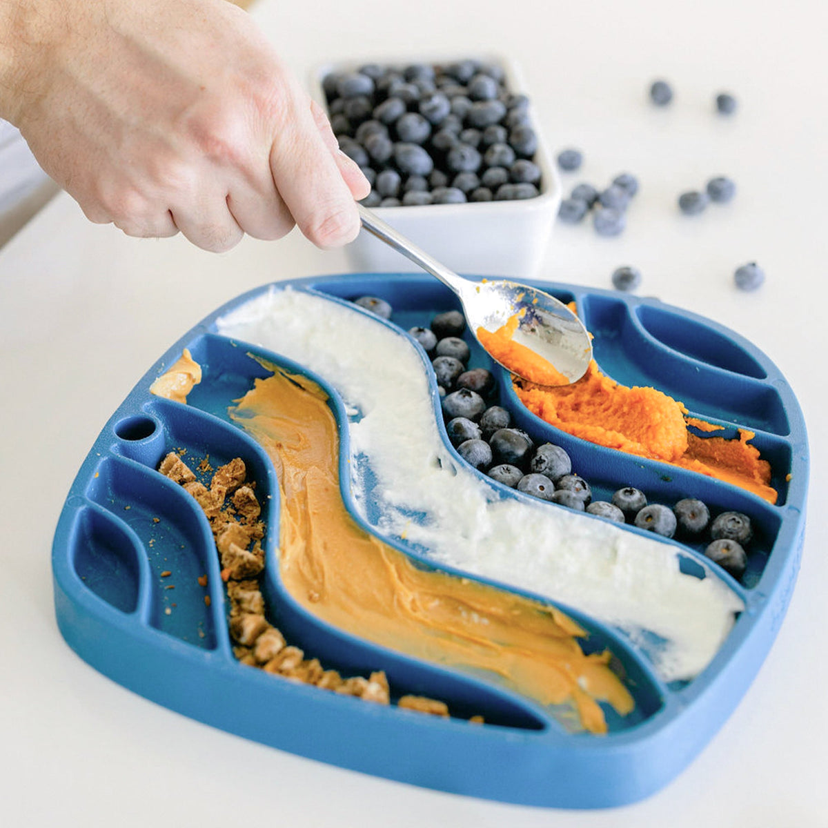 Person spreading yogurt, puree and blueberries on a blue wavy lick mat
